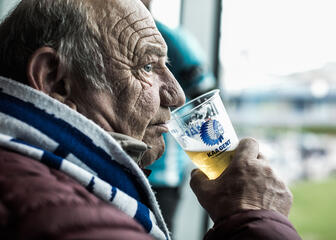 Een oude supporter drinkt een biertje uit een beker met opdruk van blauw-witte mascotte van KAA Gent.