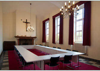 een ruimte met een open haard, rode gordijnen voor de ramen, een rood tapijt, tafels in een o-vorm met stoelen 