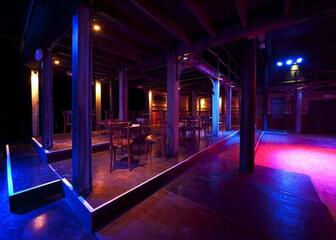 Donkere zaal met blauwpaarse verlichting, zo goed als leeg , met enkele houten tafels en stoelen. 
