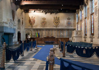 grote ruimte met wapenschilden op de muren, een open haard, een blauwe loper, blauwe stoelen, houten palen met blauwe doeken tussen