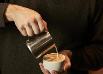 Man met bruine trui giet melk in een tas koffie