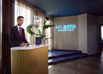 Photo of the Skybar reception, with “Mr. Sato cocktails and food” in blue neon lights on the wall and a smiling man at the reception 