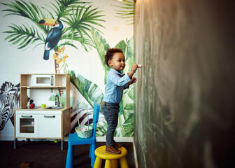 Niño pequeño subido a una silla que dibuja con tiza en una pared de pizarra 
