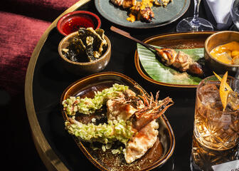 A table at a dark red sofa with some Asian dishes and 2 drinks 