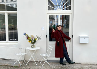 femme portant un long manteau bordeaux, posant devant une façade blanche 