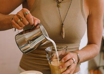Une femme avec un T-shirt beige verse du lait dans un verre rempli de café