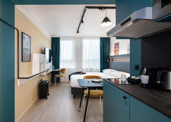 View of room with modern kitchenette, dining area, black bed and, on the left, a white hanging desk with dark yellow chair 