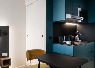 Room with seating area with black table and dark yellow chair, in the background you can see a modern kitchenette 