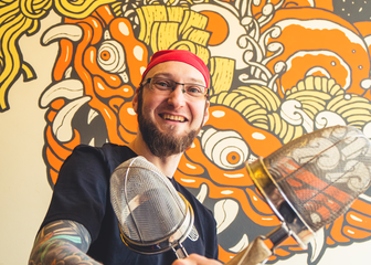•	Chef Nick is posing with crossed colanders in front of a Japanese mural
