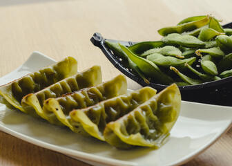 •	5 green gyoza on a white plate with edamame in a black bowl
