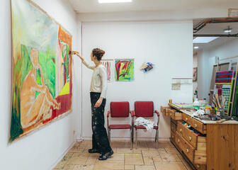 Eine Malerin arbeitet in ihrem Atelier an einem farbenfrohen Werk. Auf dem Tisch liegen Pinsel, Farben und Lappen.