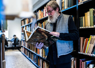 Professor Maximilian Martens browses the book 'Lam Gods - Art, History, Science and Religion' of which he is a co -author between the book racks in the University of Ghent.
