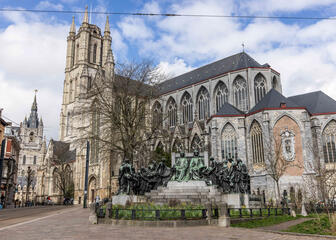 Die Statue der Brüder Van Eyck, mit einem kleinen Park davor, in dem Narzissen blühen. Im Hintergrund ist die St.-Bavo-Kathedrale und in der Ferne der Belfried zu sehen.