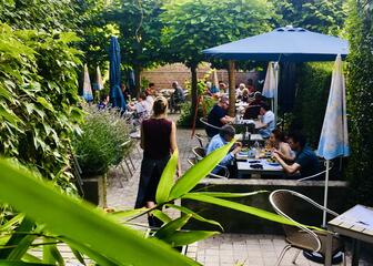 Acogedora terraza ajardinada con muchos árboles y plantas. Hay mucha gente comiendo.