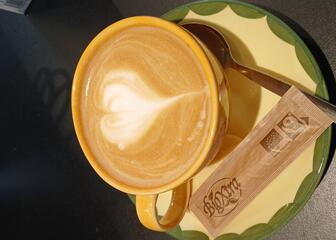 Tasse Kaffee mit Milchschaum hat die Form eines Herzens, auf dem Unterteller liegen Zucker und ein Löffel