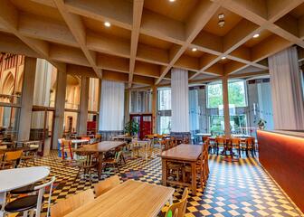 Innenansicht des Saals mit Tischen und Stühlen. Große Fensterfronten, Holzdecke mit Spots und ein Mosaikboden