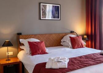 Tweepersoonsbed in een hotelkamer, met daarop 2 badjassen en rode kussens. Op de nachttafels aan beide kanten van het bed brandt een nachtlampje.