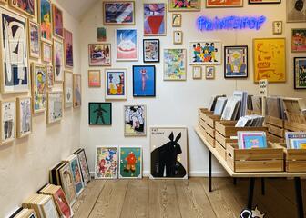 zicht in de winkel. Wand met kaders van verschillende formaten met kunstwerkjes. Tafel met houten bakken gevuld met tekeningen.