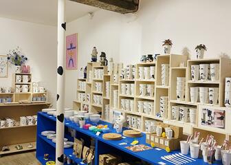 zicht in de winkel. Wand met houten rek gevuld met kopjes met daarop verschillende tekeningen. Daarvoor een blauw rek op wielen gevuld met allerlei accessoires. 