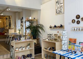 zicht in de winkel tafel met carrousel gevuld met postkaartjes, daarnaast houten kadertjes. Houten rekjes met daarop kopjes en vaasjes in keramiek