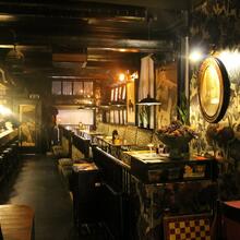 Lange ruimte met links de bar met krukken en rechts houten tafels met muurbanken en stoelen. Antiek behangpapier met grote bloemen.