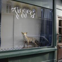 Raam met de naam "Jiggers", een opgezette vos in het raam en blauwe gordijnen. Groene gevel en een bruine deur.