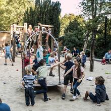 Una gran pandilla de niños juega con varios juguetes de madera y metal. La superficie es de arena. Es un día de verano.