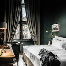 Tweepersoonskamer met kingsize bed, diepgroene muren en grijze gordijnen. Door het raam zie je een iconische gevel in de stad. 