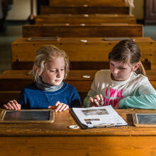 Twee schoolmeisjes in een klaslokaal aan een oude houten schoolbank met leitjes voor zich en een invulblad in het Museum De school van Toen; een rij lege bankjes is te zien achter de eerste bank.