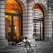 The entrance to a theatre. Two people are having a picnic on the steps at the front. A bicycle is parked at the front.