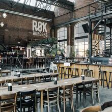 A large hall with tables and chairs, a bar with bar stools and a spiral staircase.