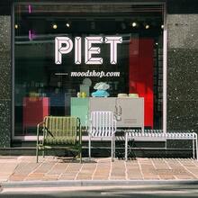 The shop window of an interior design shop. The words ‘PIET moodshop.com’ are written on the window in white letters. In front of the shop window are a few pieces of designer garden furniture.